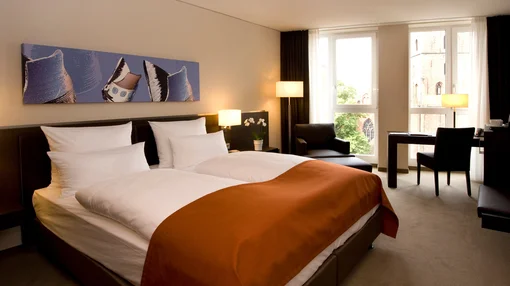 A warm and inviting bedroom with a dark wooden headboard, a pops of orange on the bedding, and a large window overlooking historic brick buildings.