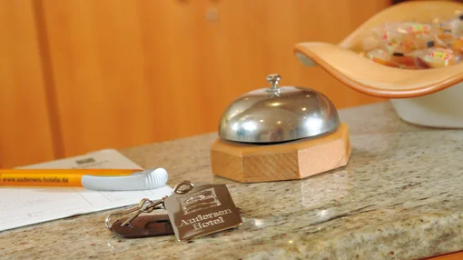 A close-up of a granite reception counter featuring a classic service bell, a hotel room key with an "Andersen Hotel" tag, and a bowl of welcome sweets.