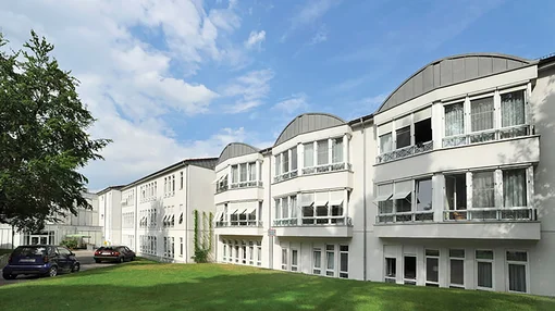 Exterior view of the modern white multi-story building at the Asklepios Clinic Birkenwerder under a blue sky, featuring large windows and curved roof elements.