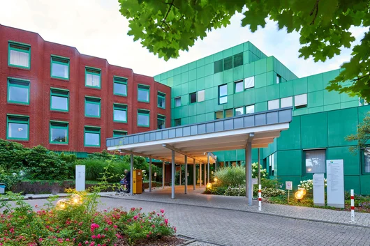 A modern multi-story medical facility with a distinctive green and red facade, featuring a covered entrance area and landscaped gardens with flowers.