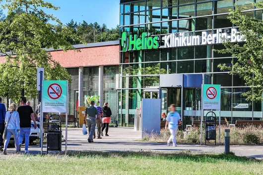 Exterior view of the modern glass entrance of the Helios Klinikum Berlin-Buch on a sunny day, with people walking towards the building and prominent "No Smoking" signs in the foreground.