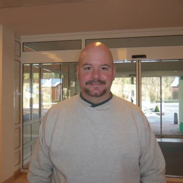 A portrait of John Wilson, a patient, standing in a lobby area with glass doors in the background.