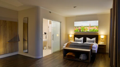 A contemporary double bedroom with dark wood flooring, an en-suite bathroom visible through a frosted glass door, and a colorful landscape painting above the bed.