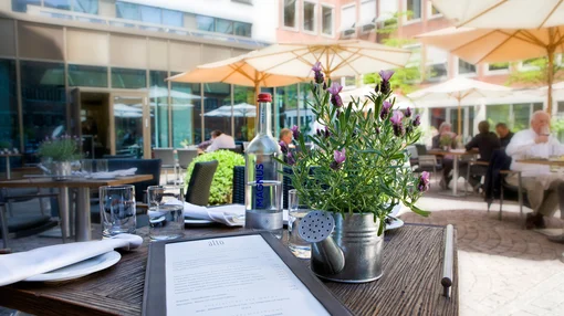 A sunlit courtyard dining area with dark wicker chairs and large umbrellas; the foreground shows a set table with a menu, bottled water, and a lavender plant.
