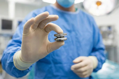 A surgeon in scrubs holding an artificial spinal disc replacement implant in the operating room.