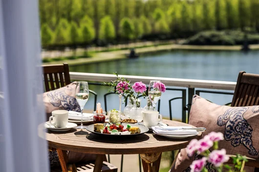 A set wooden table with breakfast items, pastries, and flowers on a terrace overlooking a serene lake and green parkland.