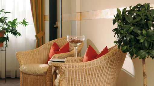 Two cozy wicker armchairs with red cushions next to a small glass table and a potted laurel tree in a warm, sunlit hotel room.