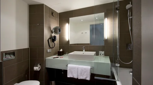 A sleek bathroom featuring dark brown wall tiles, a wide white vessel sink on a frosted glass counter, and a walk-in glass shower partition.