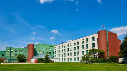 A wide shot of the modern, multi-story white and green clinic building under a bright blue sky, featuring a large green lawn in the foreground.
