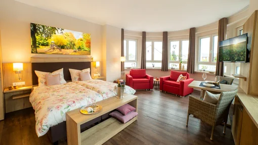 A large corner suite with multiple windows, two red armchairs, a dining area, and a plush double bed decorated with floral patterns and a champagne bucket on a bench.