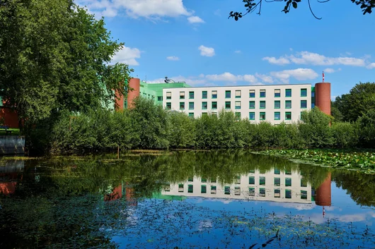 A scenic view of the clinic building reflected in a calm pond with lily pads, framed by green trees under a clear sky.