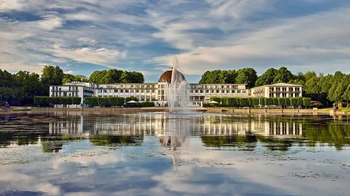 Exterior View of Parkhotel Bremen by the Lake A wide-angle shot of the historic Parkhotel Bremen building reflected in a calm lake with a central fountain under a blue sky.