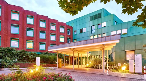 A warm evening shot of the clinic's covered entrance area, featuring glowing spherical lights, flower beds, and the distinctive red brick and green facade.