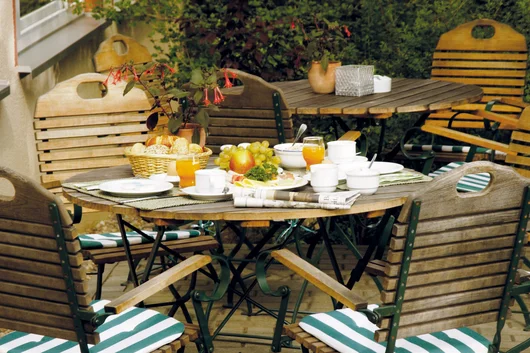 A rustic wooden garden table set for breakfast with fresh fruit, juice, and coffee in a lush, green outdoor courtyard.