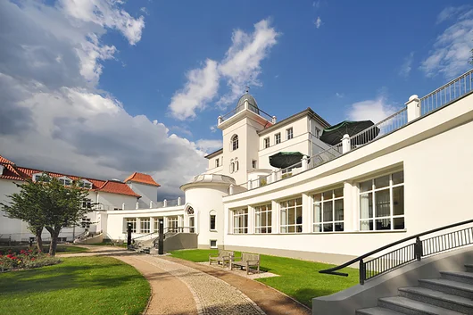 Low-angle exterior shot of the white historic building at Asklepios Clinic Birkenwerder, featuring a sunny courtyard with green lawns, stone paths, and a terraced balcony.
