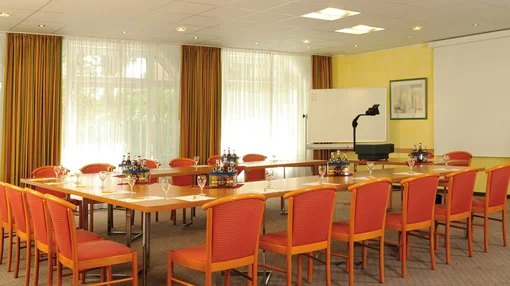 A large, U-shaped wooden conference table with orange chairs, set with water bottles and glasses, in a bright room equipped with a whiteboard and projector screen.