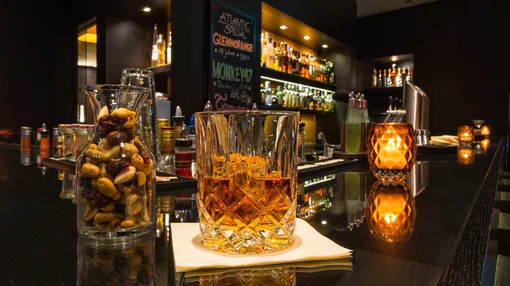 A close-up of a crystal whiskey glass and a jar of mixed nuts on a polished black bar counter, with warm candlelight and a chalkboard menu in the background.