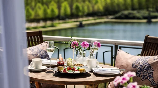 Breakfast with a View at Parkhotel Bremen A wooden table set for two with pastries, coffee, and wine on a balcony overlooking a scenic lake and lush green trees.