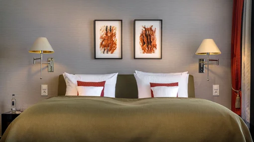 Superior Double Bed Detail A close-up of a neatly made large double bed with green bedding, white pillows with red accents, and modern wall lamps on a neutral background.