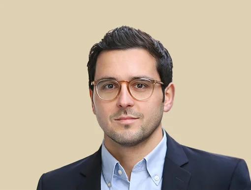 Headshot of Dr. Nils Farhan with short dark hair, wearing glasses, a light blue button-down shirt, and a dark blazer, set against a solid beige background.