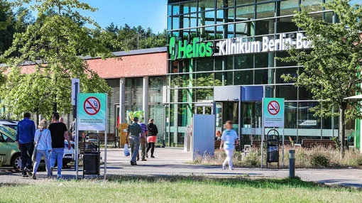 Exterior view of the modern glass entrance of the Helios Klinikum Berlin-Buch on a sunny day, with people walking towards the building and prominent "No Smoking" signs in the foreground.