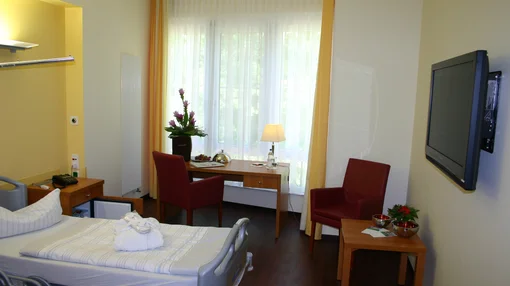 Interior of a high-end hospital room at Asklepios Clinic Birkenwerder, featuring a modern medical bed, wooden furniture, red armchairs, a wall-mounted TV, and bright natural light from a large window.