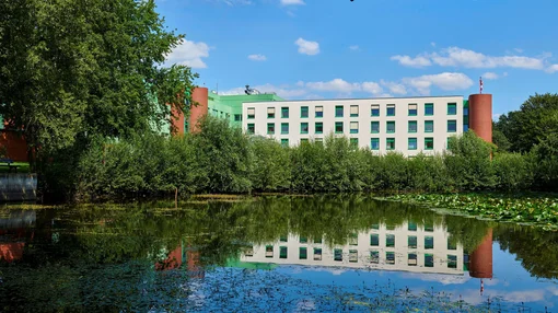 A scenic view of the clinic building reflected in a calm pond with lily pads, framed by green trees under a clear sky.