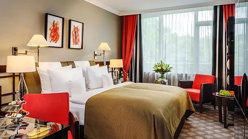 Comfortable Classic Double Room A cozy hotel room with a large double bed, red accent chairs, a small dining table with treats, and large windows providing natural light.