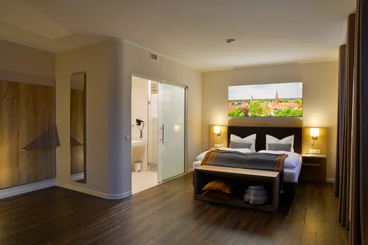 A spacious, contemporary hotel room with a large bed, wooden floors, and an en-suite bathroom with a glass sliding door.