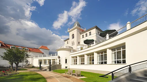 Low-angle exterior shot of the white historic building at Asklepios Clinic Birkenwerder, featuring a sunny courtyard with green lawns, stone paths, and a terraced balcony.