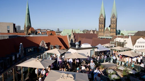 A wide shot of a busy rooftop event at the Atlantic Grand Hotel, with guests enjoying drinks under umbrellas and the spires of the Bremen Cathedral in the background.