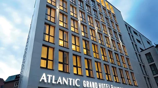A low-angle exterior shot of the contemporary stone building of the Atlantic Grand Hotel Bremen at dusk, featuring large glass windows and glowing hotel signage.