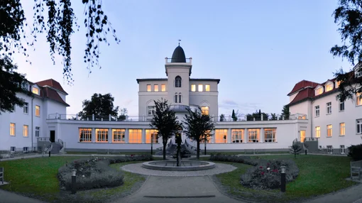 Wide panoramic night view of the illuminated historic main building of the Asklepios Clinic Birkenwerder, showing the central tower and glowing windows reflected in the courtyard.